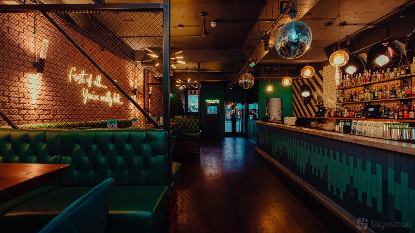 A cocktail bar with teal leather booths, a patterned bar counter, disco balls, and neon signage at Tequila Mockingbird Wimbledon.