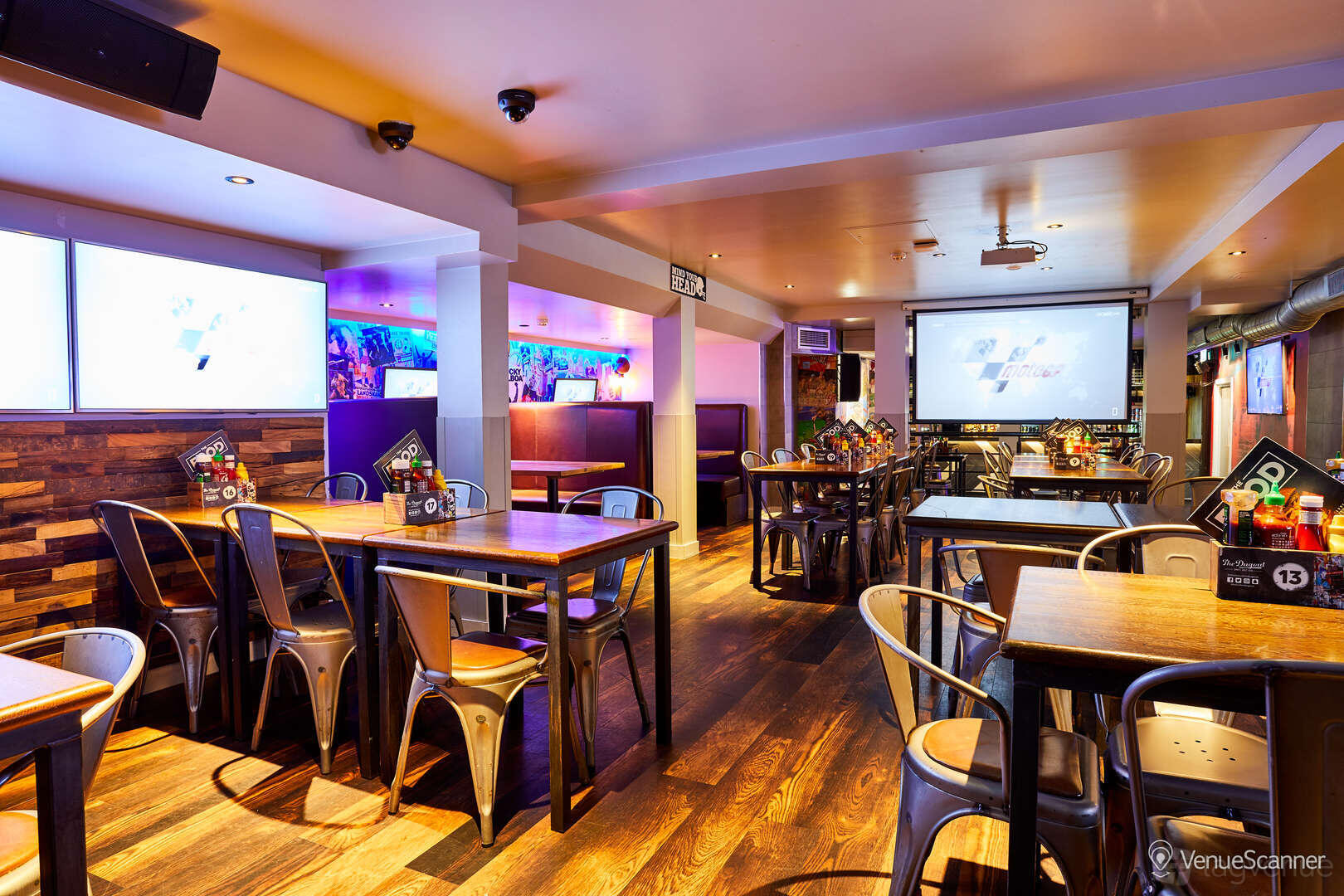 An event space in a bar with wooden floors, metal chairs, wall screens, and sports decor at Belushi's & The Dugout London Bridge.