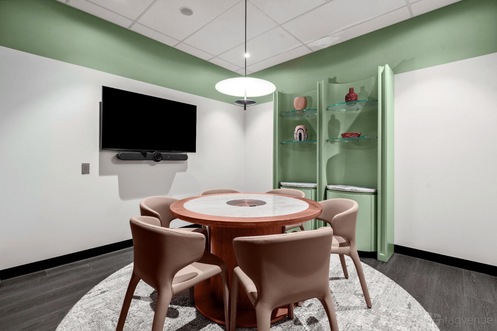 A meeting room with a round table, four beige chairs, wall-mounted TV, and built-in green shelving at The Great Room.