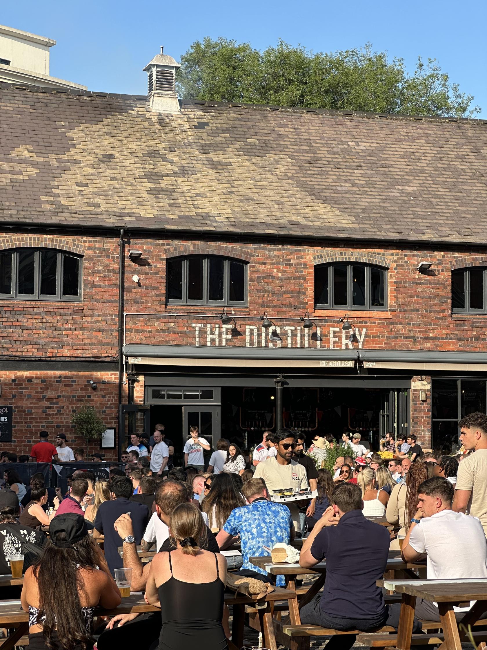 A pub with a red brick exterior, arched windows, and a crowd gathered on a sunny patio at The Distillery.