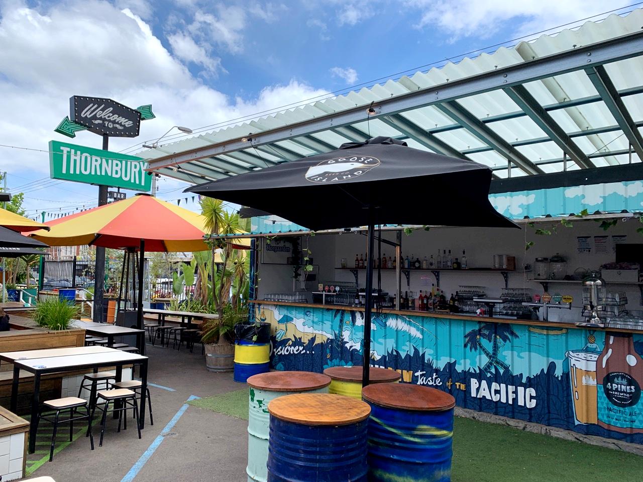 An outdoor restaurant space with shaded barrel tables, colorful umbrellas, and a container-style bar at Welcome To Thornbury.
