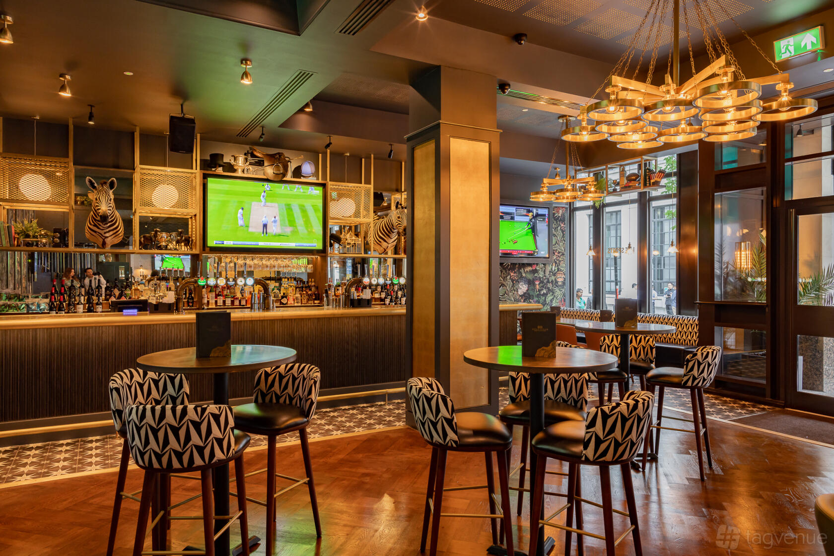 A lounge in a pub with patterned chairs, round tables, and large TVs displaying sports at Goldwood.