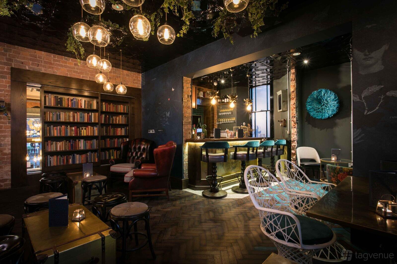 A bar in a restaurant with a gold-lit counter, velvet chairs, bookshelves, and hanging glass lights at The Lost and Found Knutsford.
