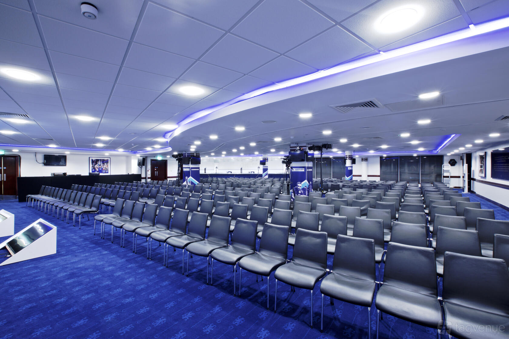 An auditorium-style stadium suite with rows of black chairs, blue carpet, and purple LED ceiling lights at Chelsea Football Club.
