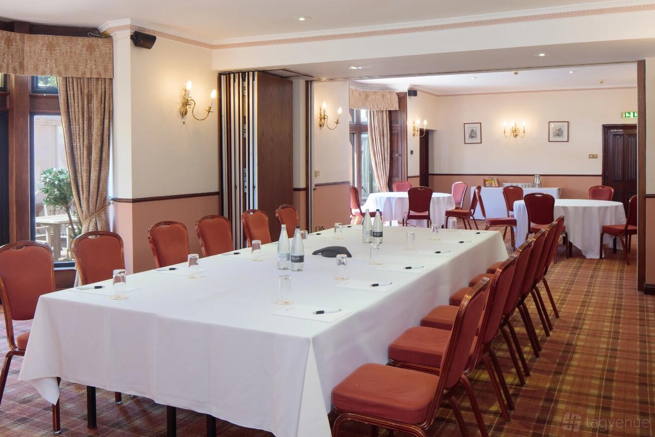 A hotel terrace room with U-shaped tables covered in white linens, red chairs, and large windows at Nailcote Hall Hotel.