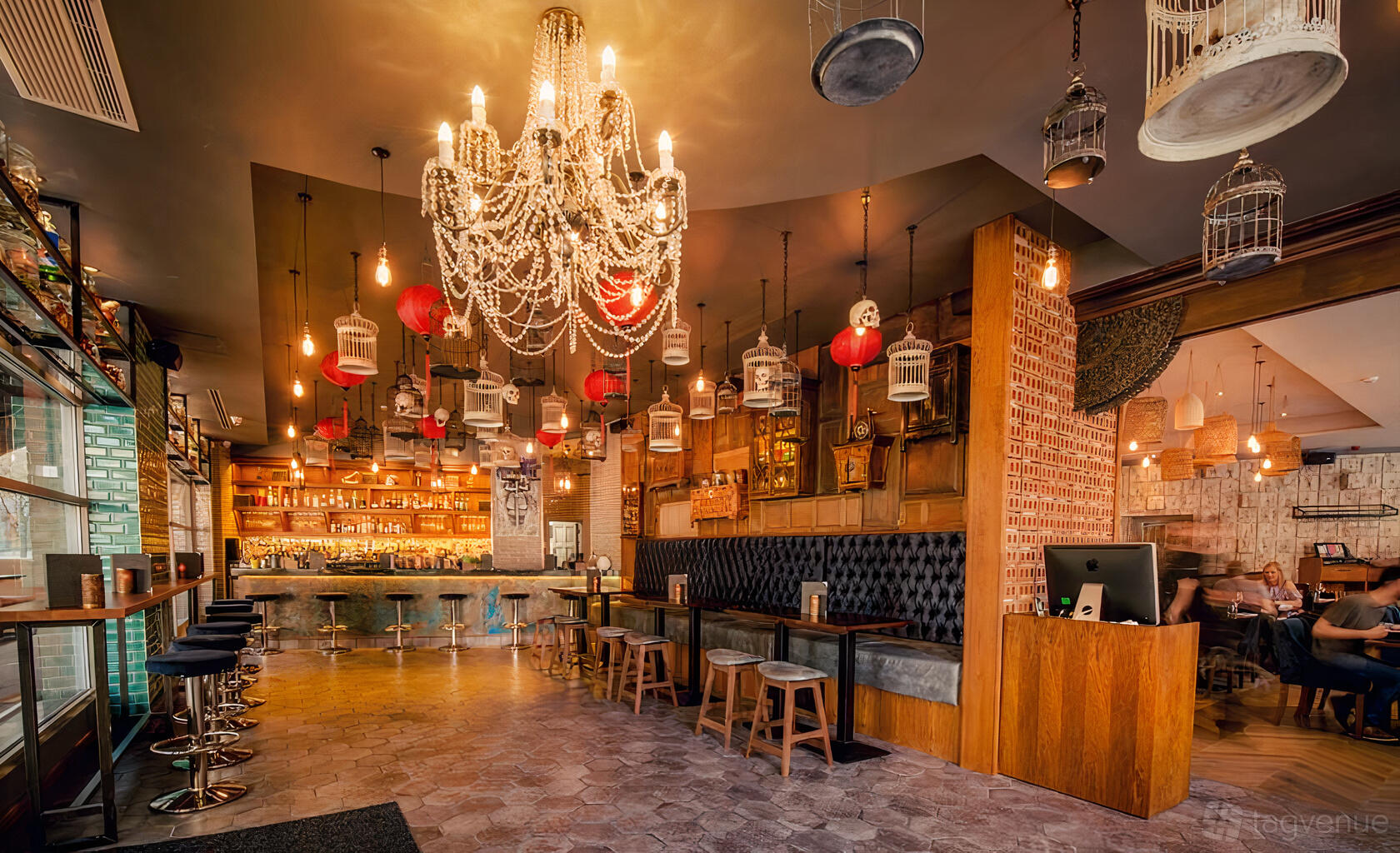 A restaurant bar area with a crystal chandelier, birdcage decor, tufted banquette seating, and a lit bar at Siamais.