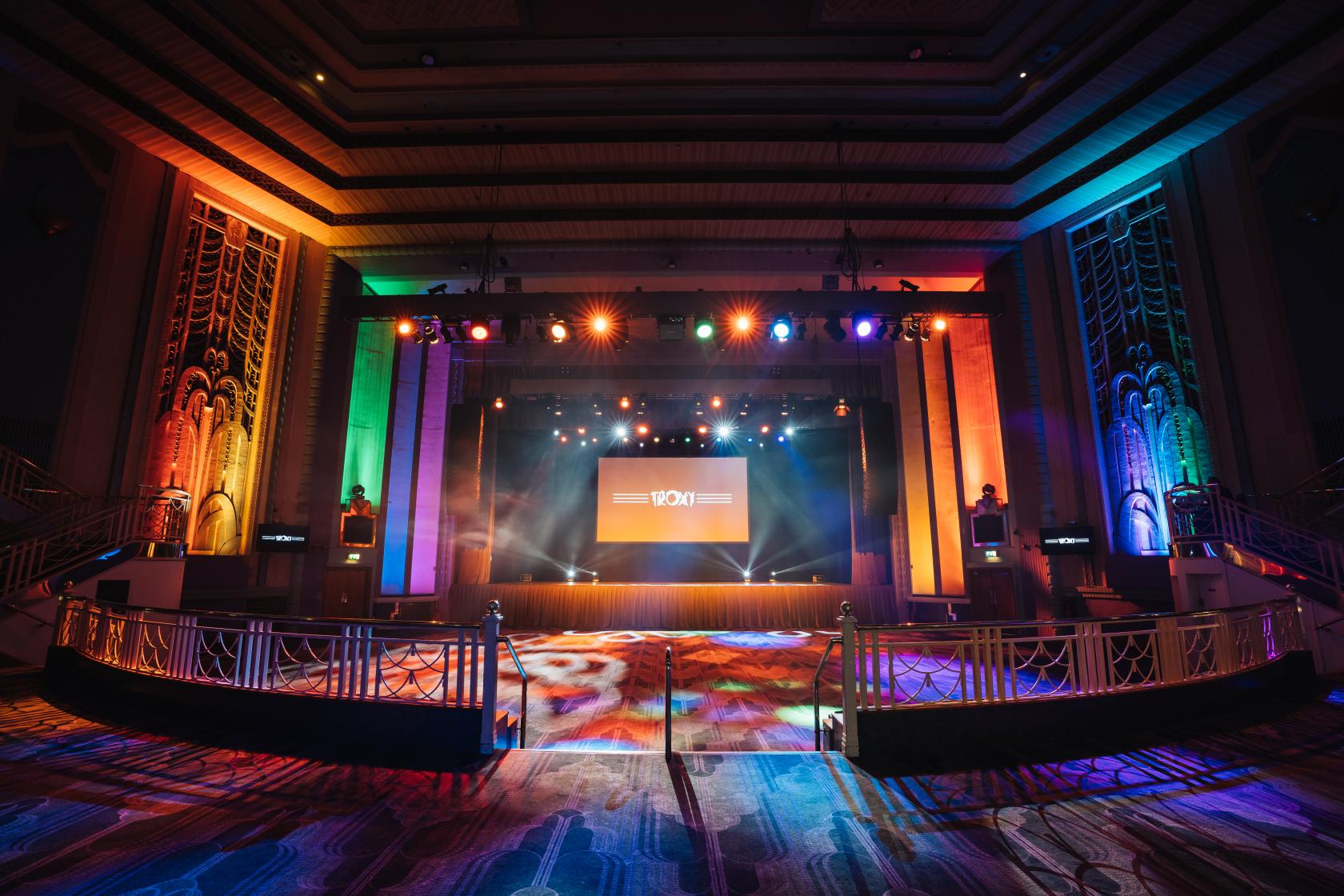 An art deco event hall with multicolored stage lighting, large central screen, and patterned carpet at Troxy.