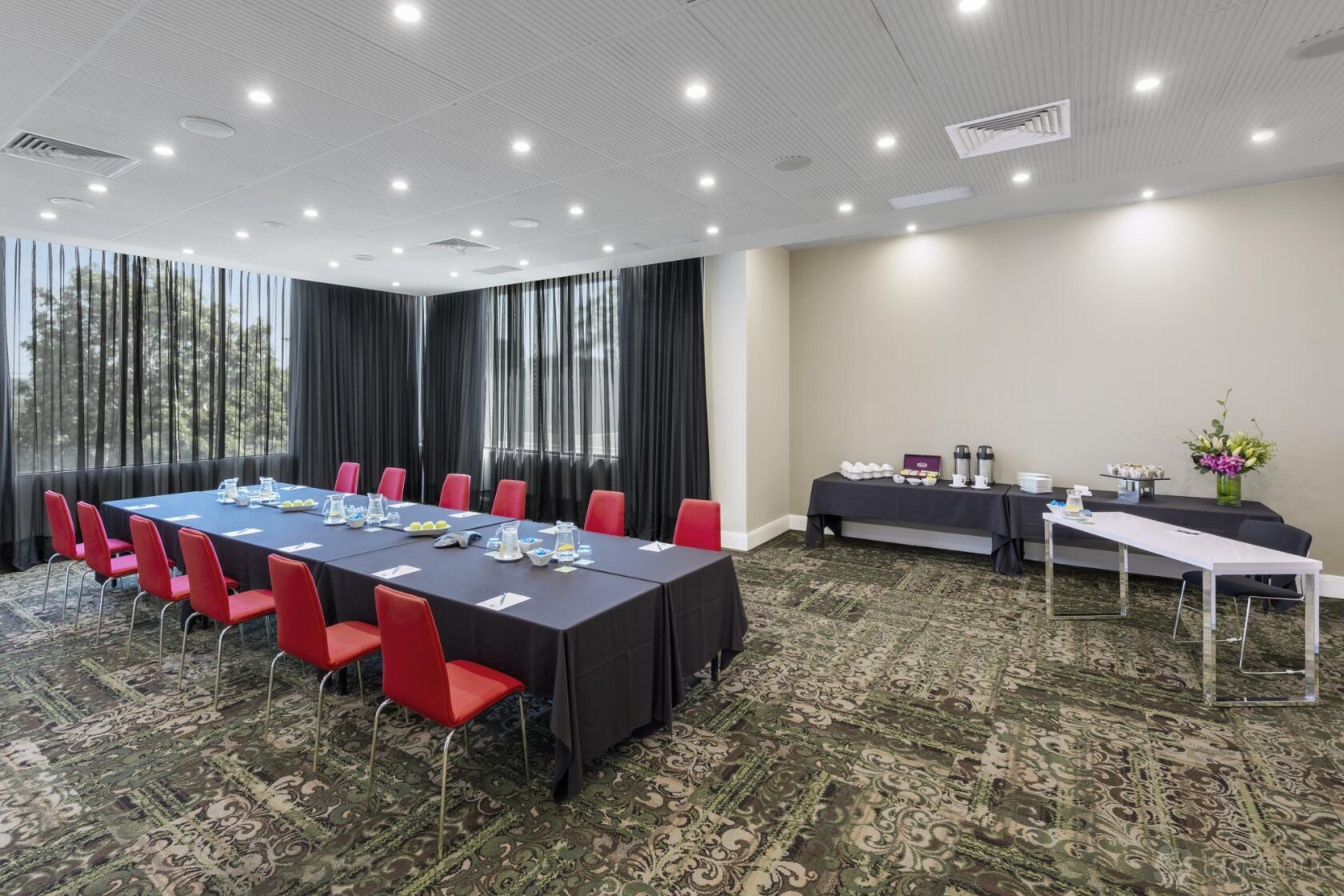 A meeting room with red chairs, dark tablecloths, floor-to-ceiling windows, and a refreshment station at Nesuto Parramatta Apartment Hotel.