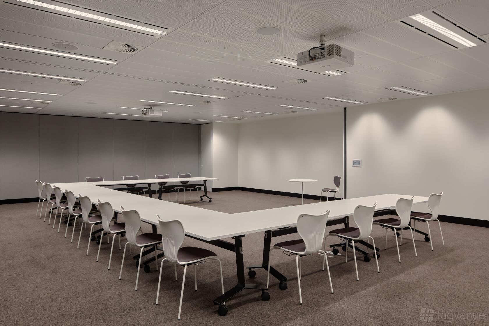 A meeting room with U-shaped white tables, ergonomic chairs, and ceiling-mounted projector at Flex by ISPT at 4 National Circuit.