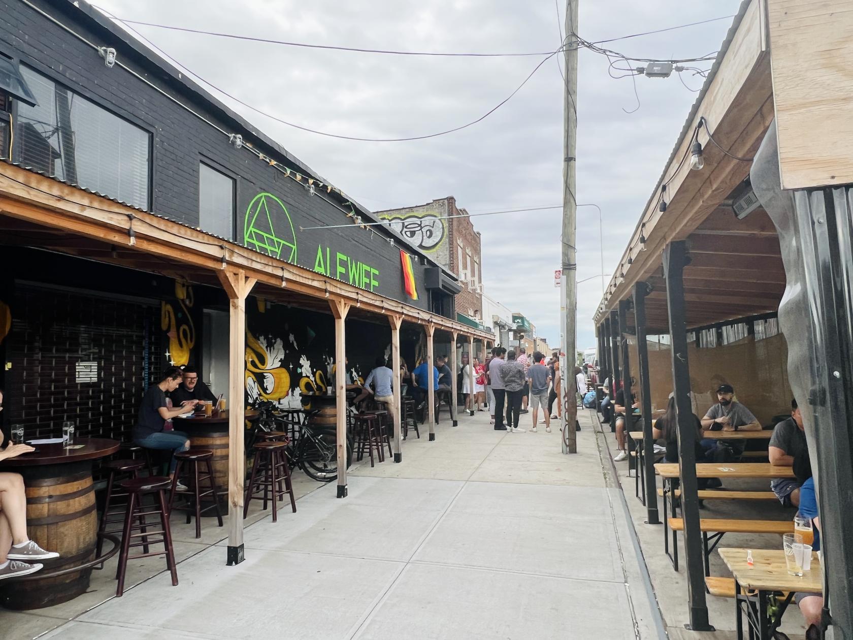 An outdoor bar with covered patio seating, wooden benches, and barrel tables at Alewife Brewing.