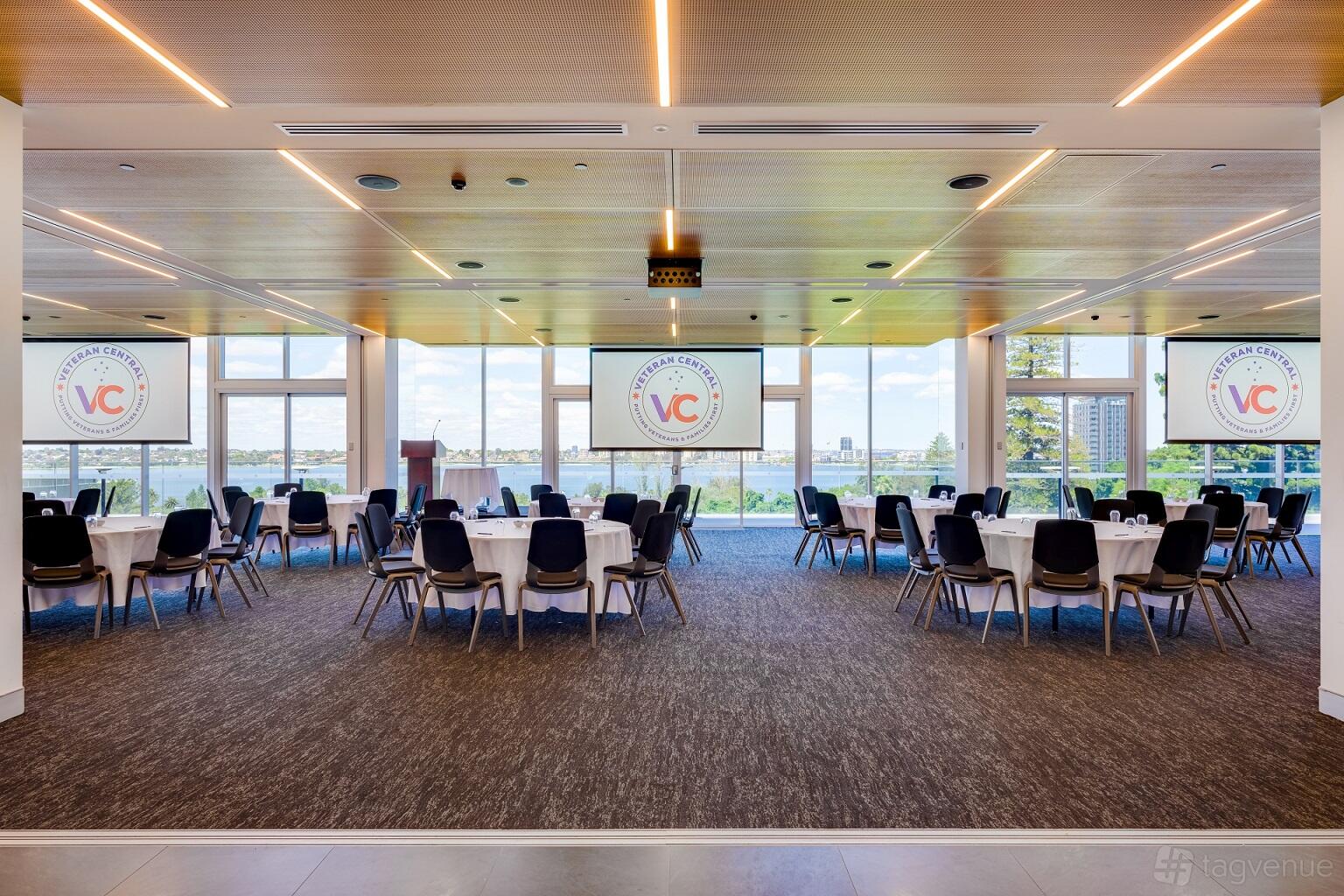 A function room with large windows, round tables with chairs, and dual projector screens at The Anzac Club WA.