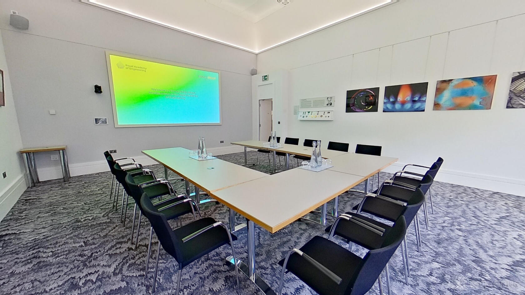 An event venue with U-shaped tables, black chairs, a projector screen, and abstract art at Prince Philip House.