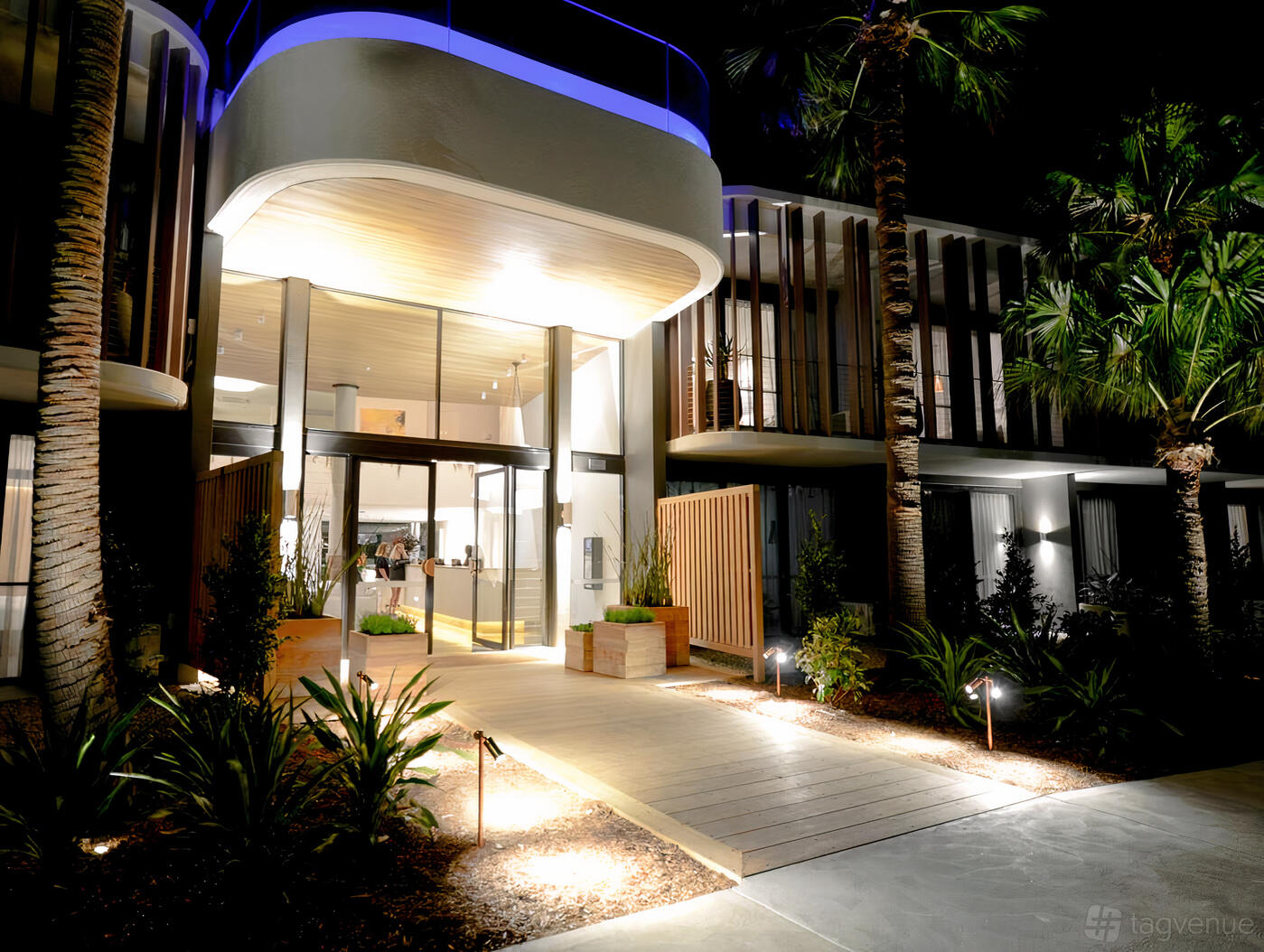 A hotel entrance with a wooden walkway, glass doors, uplighting, and surrounding palm trees at Bannisters Pavillion.