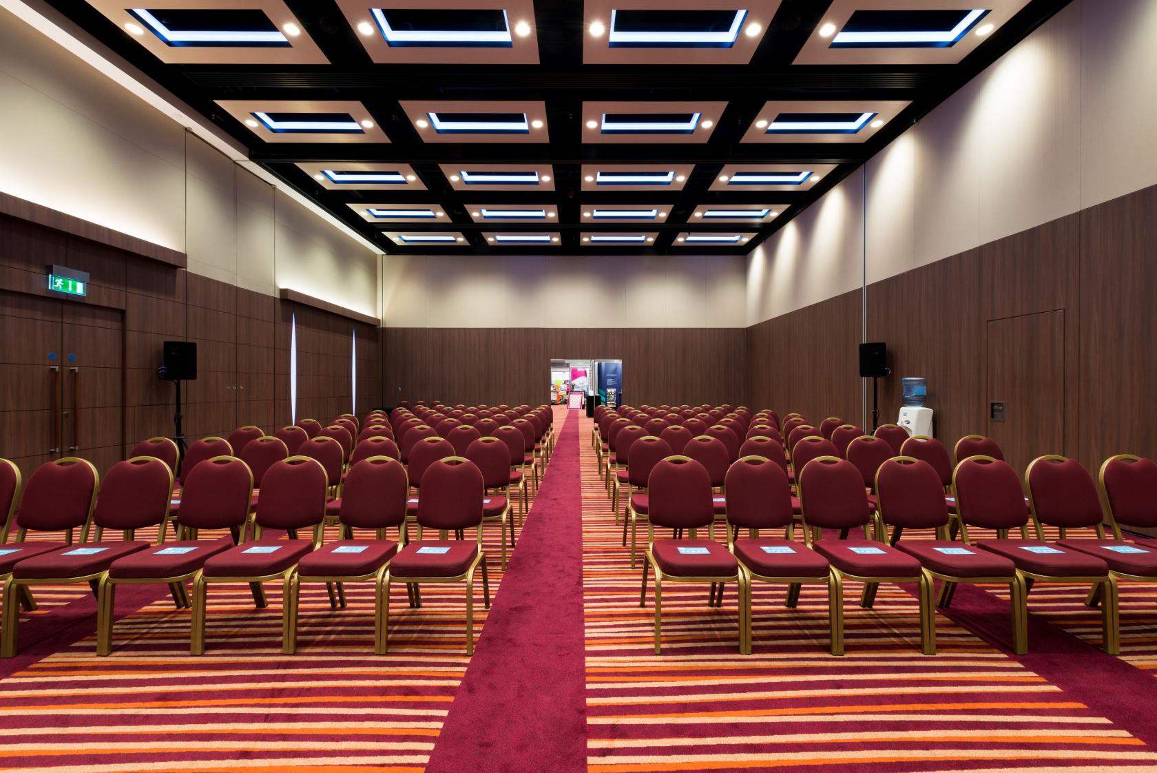A hotel meeting room with rows of red cushioned chairs, striped carpet, and geometric ceiling lights at Mercure London Earl's Court.