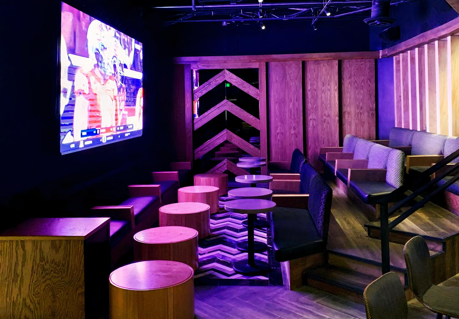 A dining room with stadium-style seating, round wooden tables, and a large projector screen at 3rd Base LA.