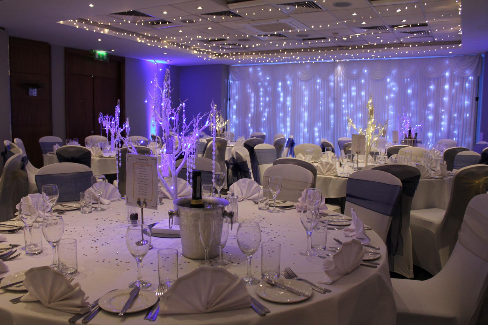 An event space with round tables set with white linens, chair covers, and LED-lit backdrop at Holiday Inn London Bexley.