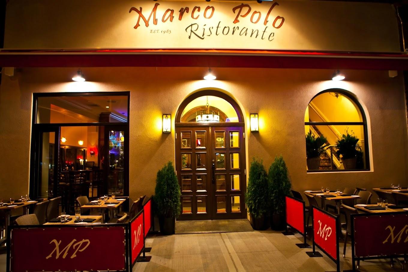 A restaurant with a warmly lit entrance, outdoor seating, and red barriers at Marco Polo.