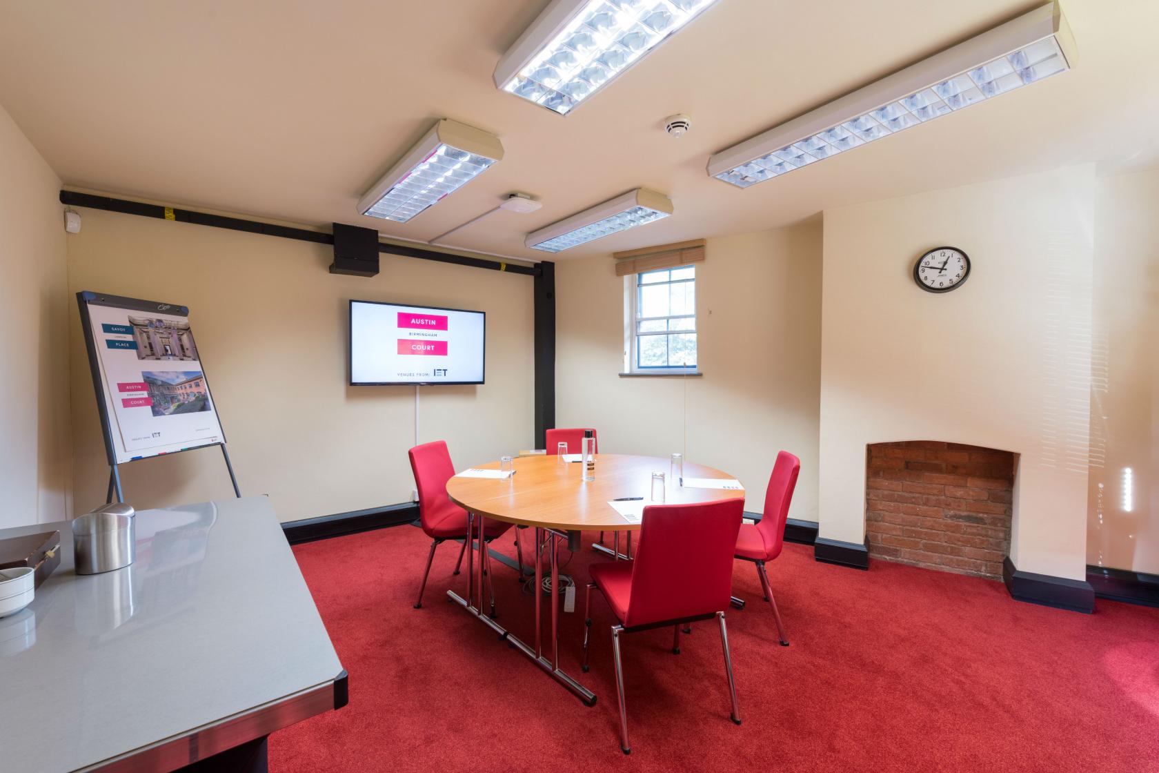 A conference room with a round table, red chairs, wall-mounted screen, and flip chart at IET Birmingham: Austin Court.