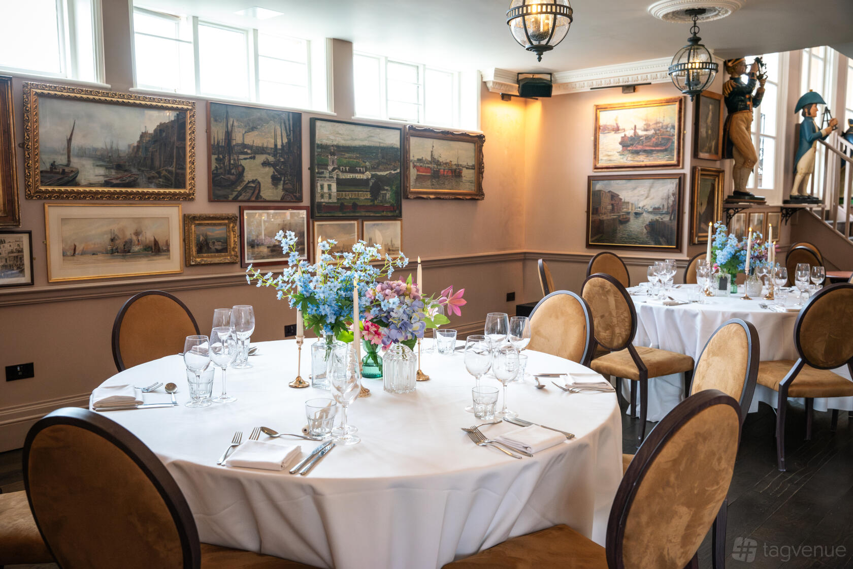 An event space in a pub with large windows, round tables set with glassware, and maritime paintings at Trafalgar Tavern.