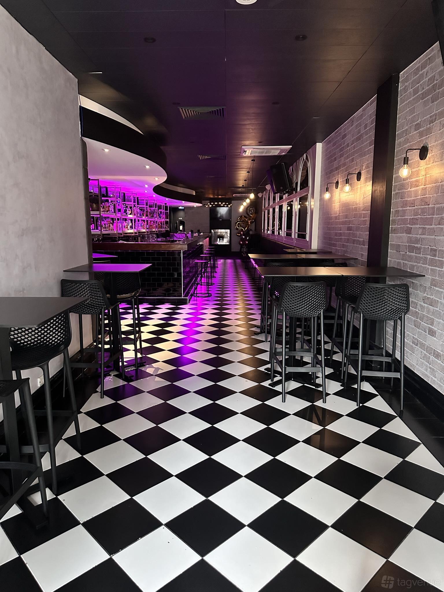 A bar with black and white checkered floor, purple accent lighting, high tables, and exposed brick at Amani Bar.