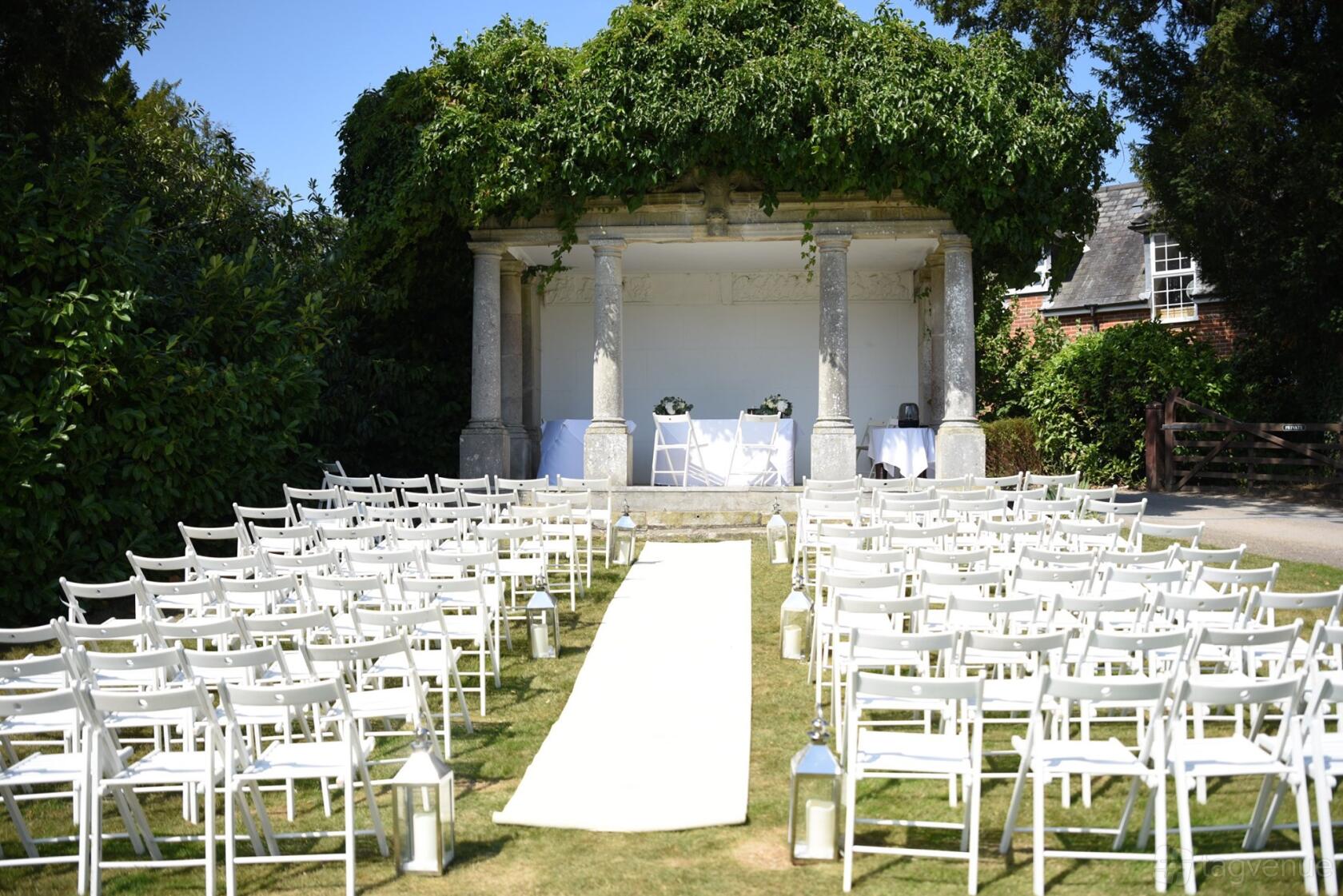 An outdoor ceremony space with white folding chairs and a vine-covered stone pavilion at Highfield Park.