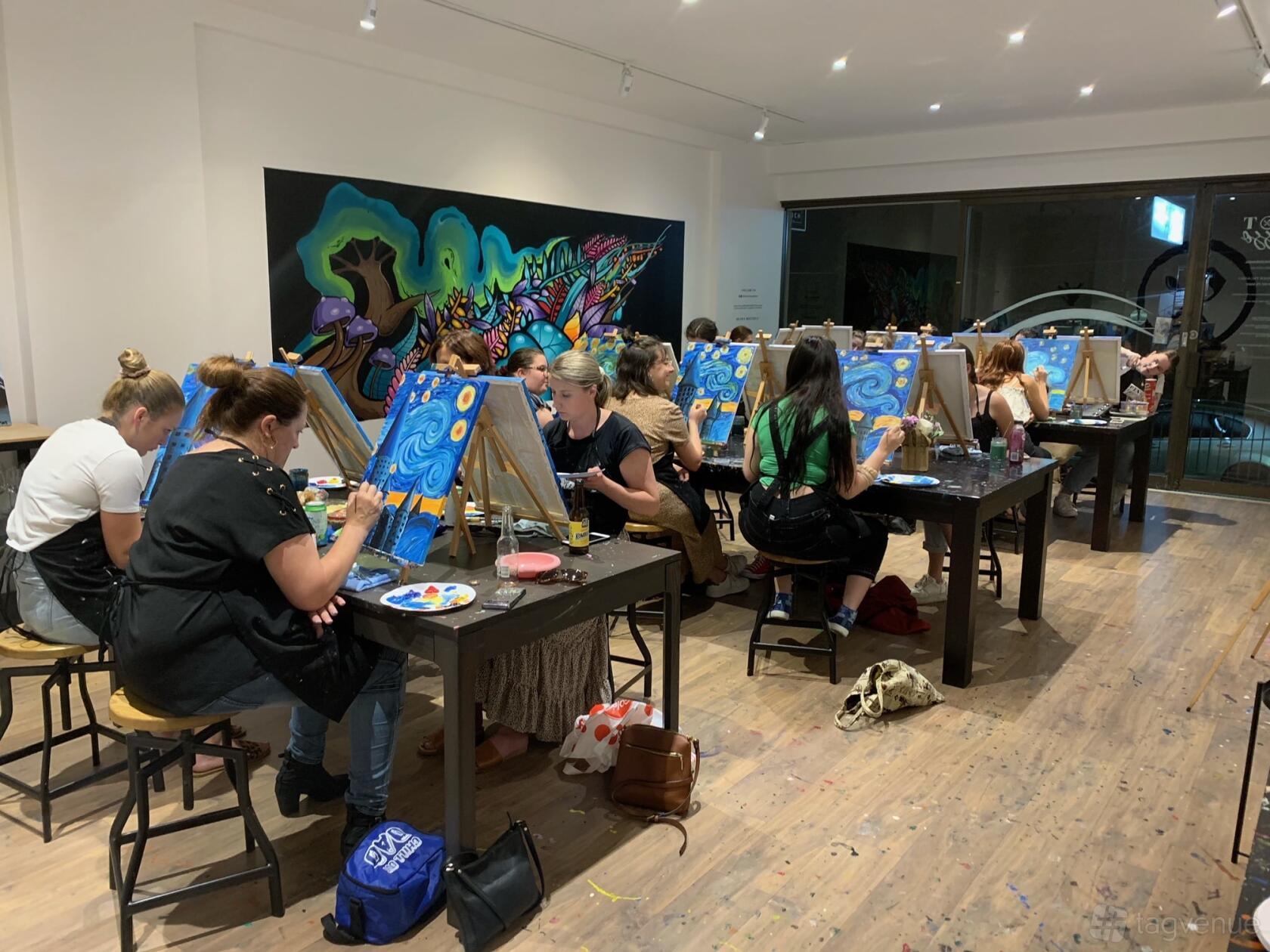 An art studio with guests painting at easels on wooden floors and a colorful mural wall at Pinot & Picasso Miranda.