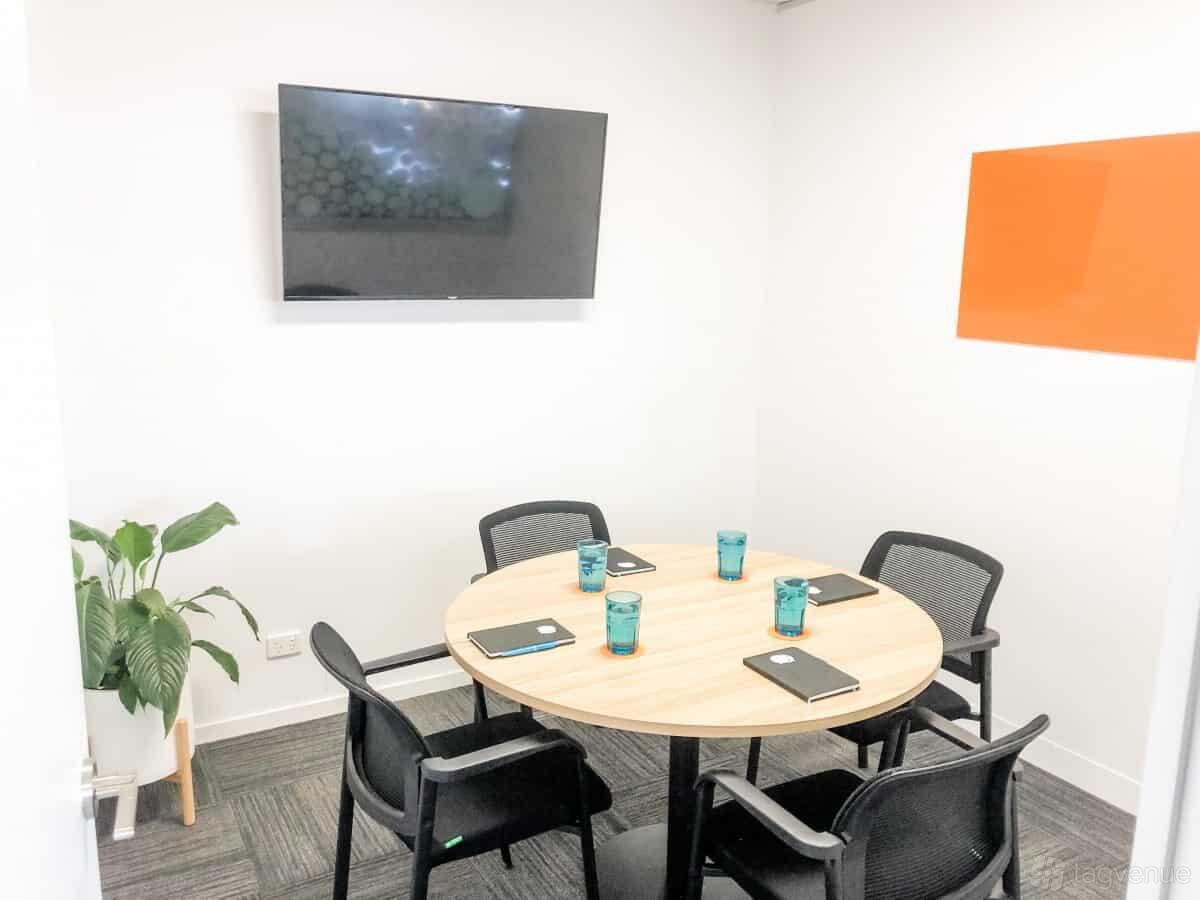 A coworking meeting room with a round wooden table, four mesh chairs, wall-mounted TV, and a potted plant at Cowork + Create.