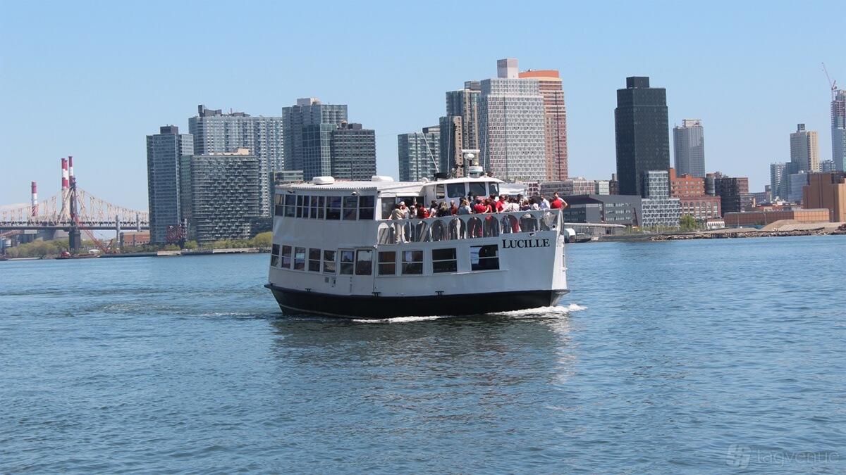 A boat with an open upper deck carrying passengers on the water with city skyline views at Empire Cruises Lucille.
