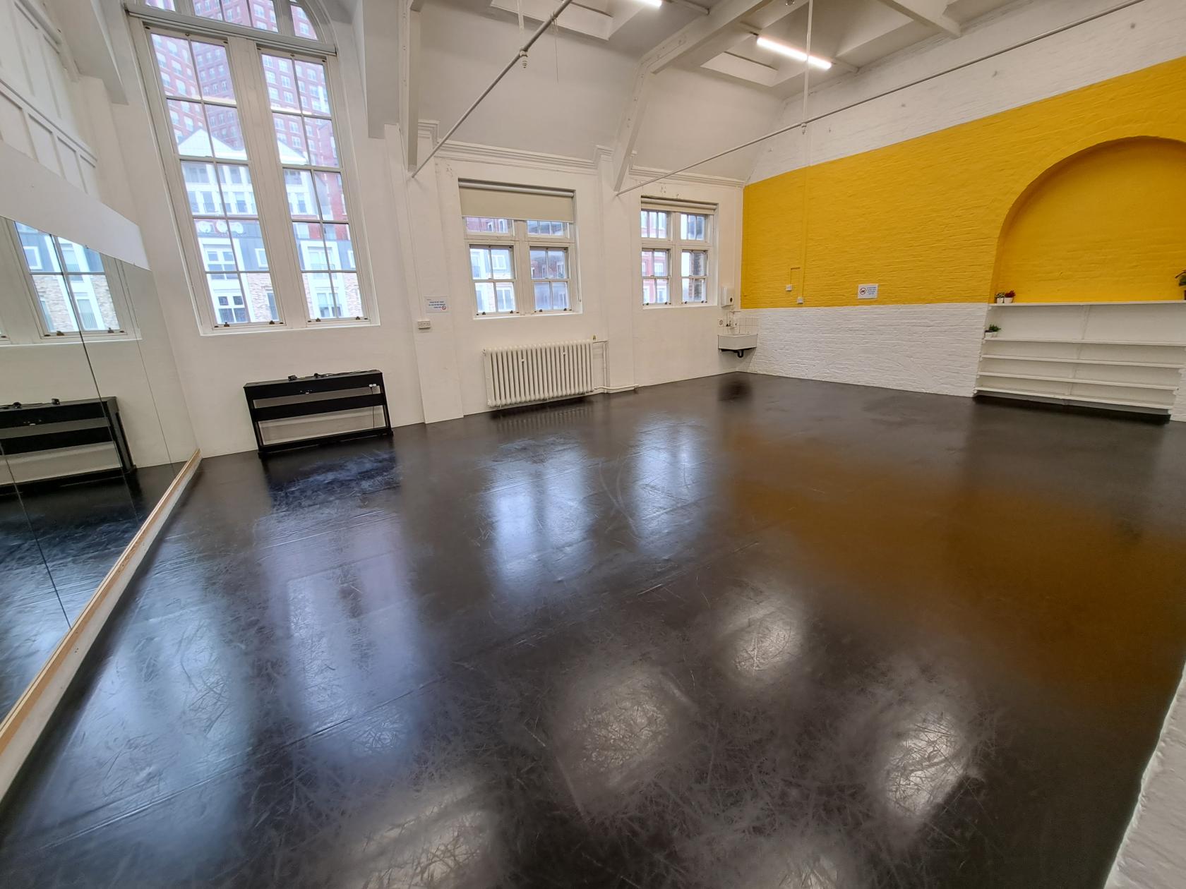 A rehearsal room with black sprung floors, large windows, a wall mirror, and a yellow accent wall at The Boury Studios.
