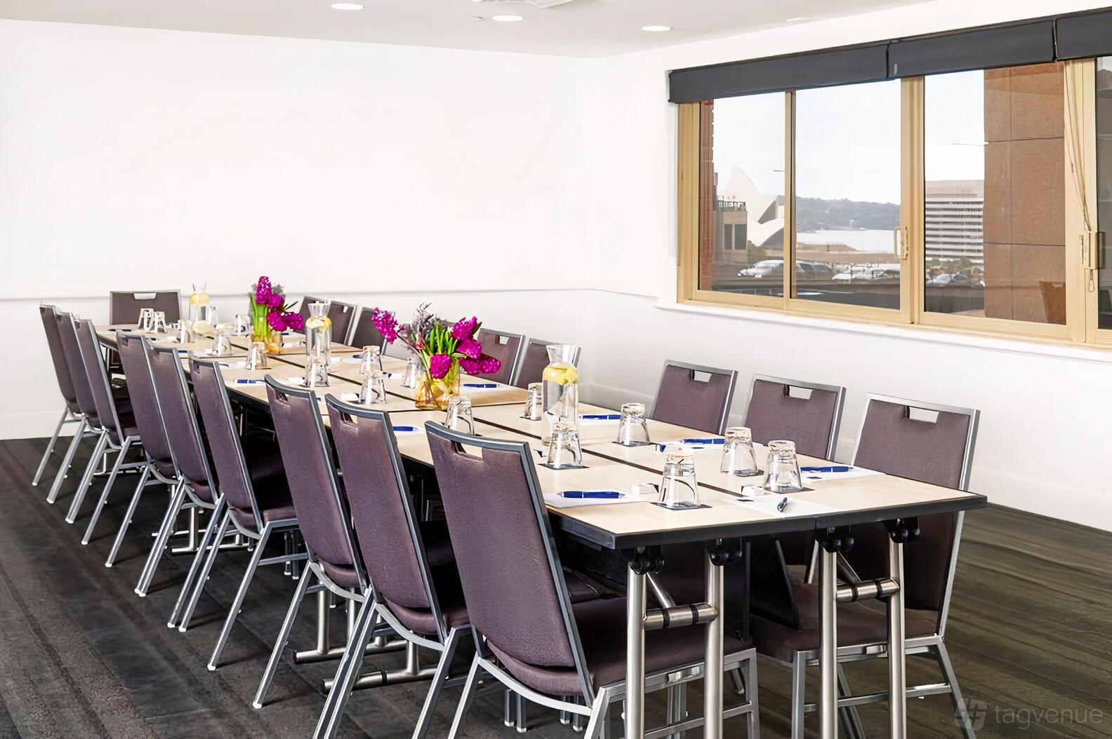 An event space with two conference tables arranged in a U-shape, purple chairs, and large windows at Sebel Quay West Suites Sydney.