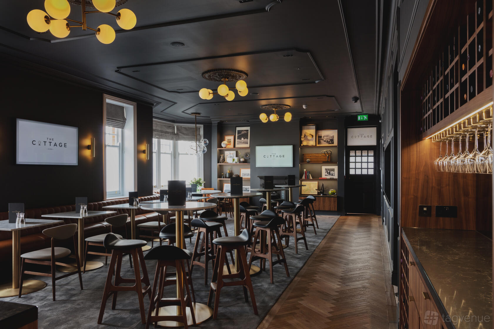 A stadium lounge with round tables, bar stools, wall shelves, and large windows at Fulham Football Club - Cottage.