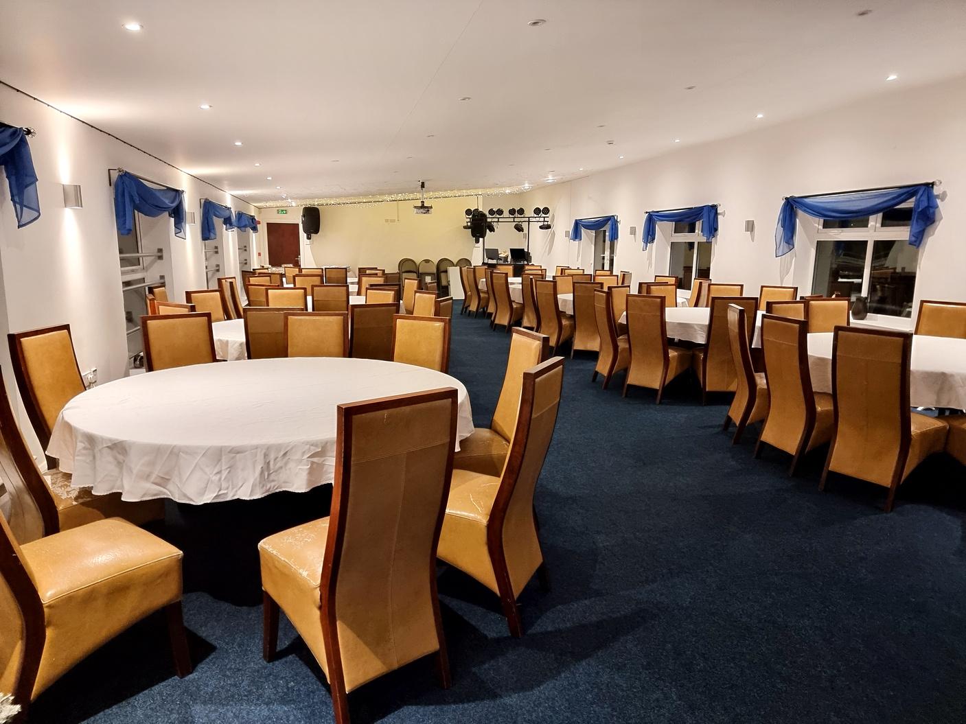 An event space with round and rectangular tables, tan chairs, and blue curtains at Leicester Road Stadium.