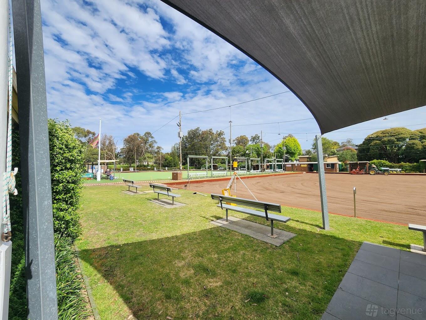 A sports club with lawn bowling greens, bench seating, and a shaded awning at Blackburn Bowls Club.