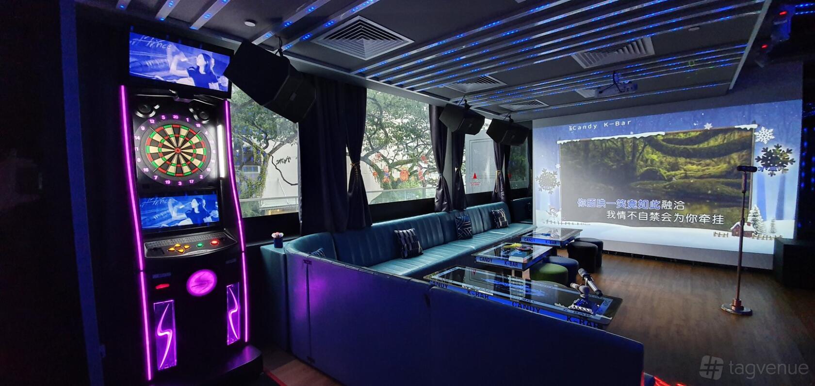 A karaoke bar with plush blue sofas, a dart machine, glass tables, and a large projection screen at Candy K-Bar Family Karaoke.