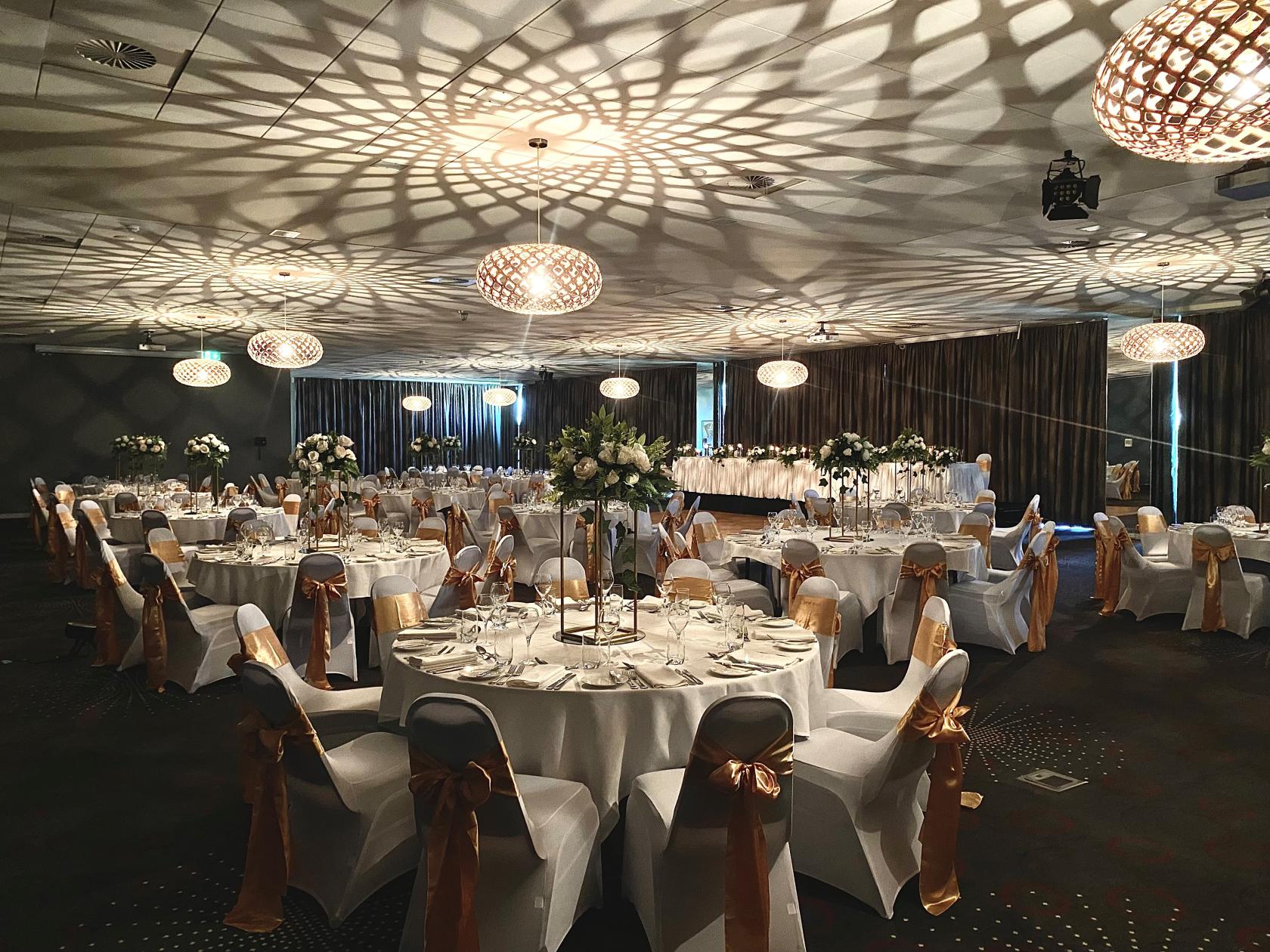 An event venue with round banquet tables, white linens, and gold chair sashes under patterned pendant lights at Melton Entertainment Park.