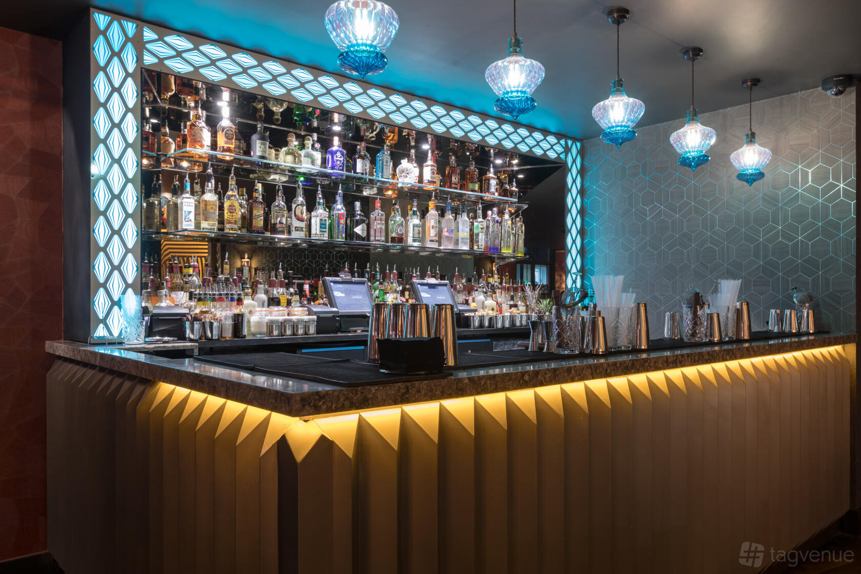 A cocktail bar lounge with an illuminated bar, mirrored bottle display, and geometric pendant lights at Dirty Martini Leeds.