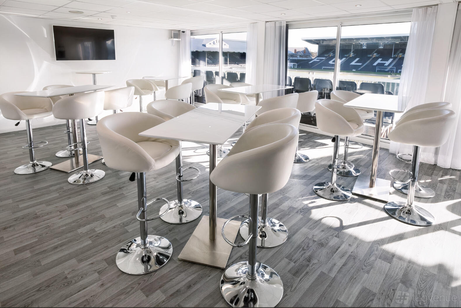 A meeting centre with white bar stools, high tables, and large windows overlooking the football pitch at Fulham Football Club.