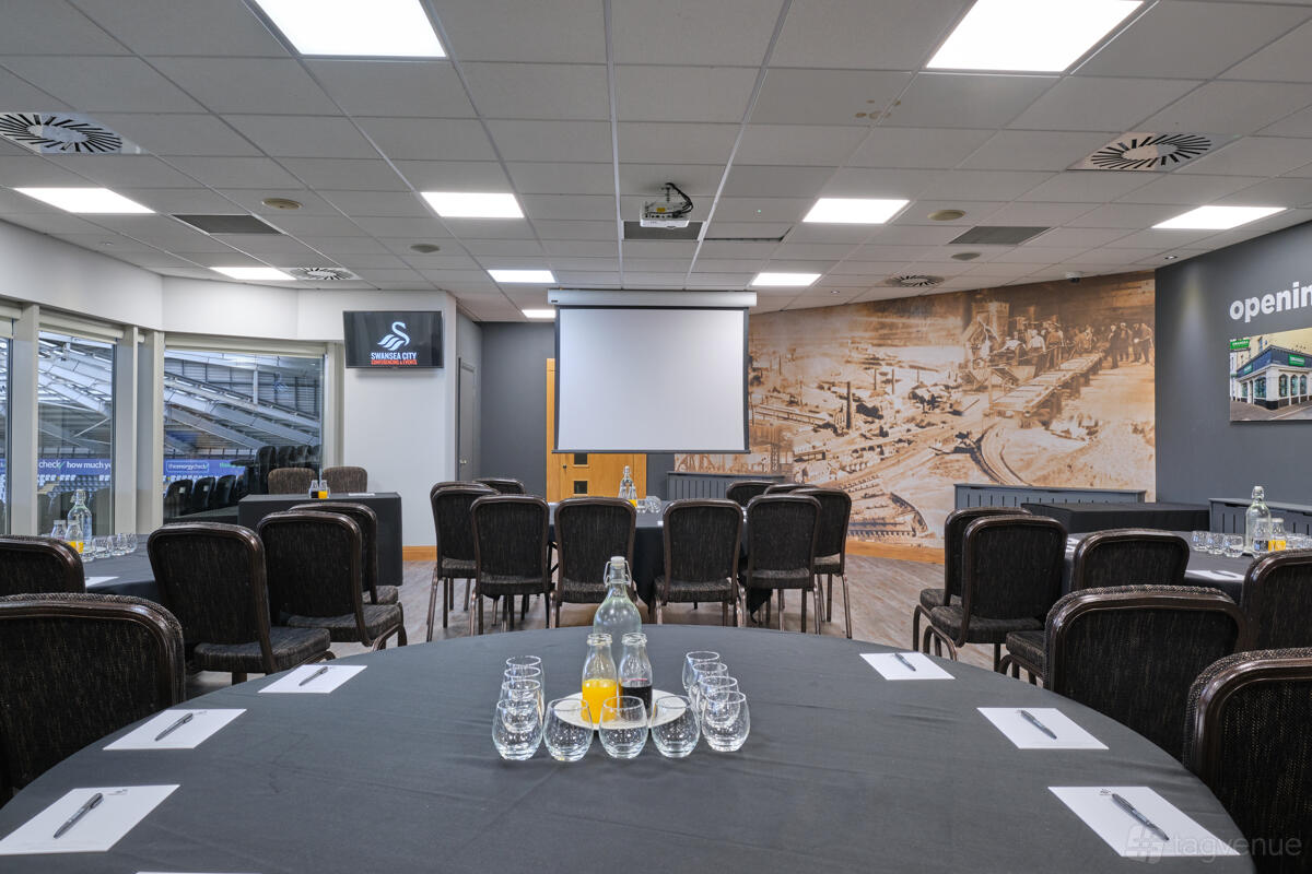 A stadium lounge with conference seating, a projection screen, and city mural walls at Swansea.com Stadium.
