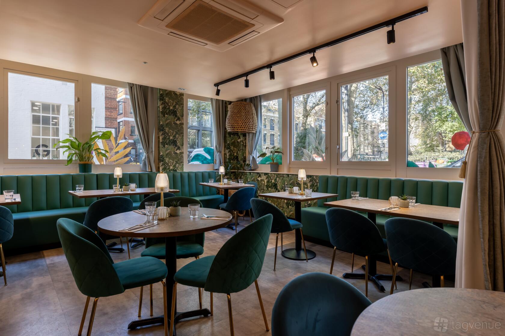 A function room in a restaurant with green velvet banquettes, round tables, and large windows at Cottons Shoreditch.