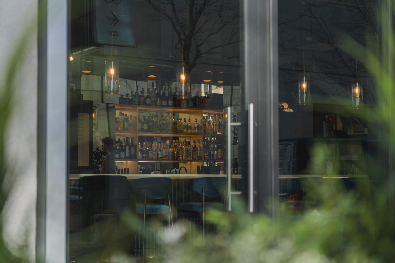 A pub with a backlit bar lined with bottles, pendant lighting, and high-backed bar stools at Qube East - Cocktail Bar & Recording Studios.