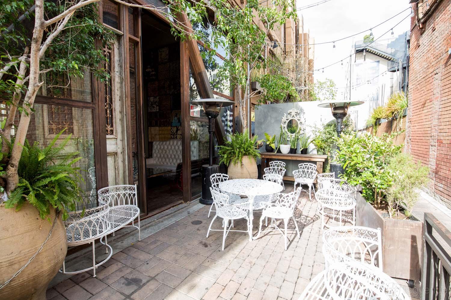 An outdoor terrace with white metal tables and chairs, potted plants, and brick flooring at Saint Felix Hollywood.