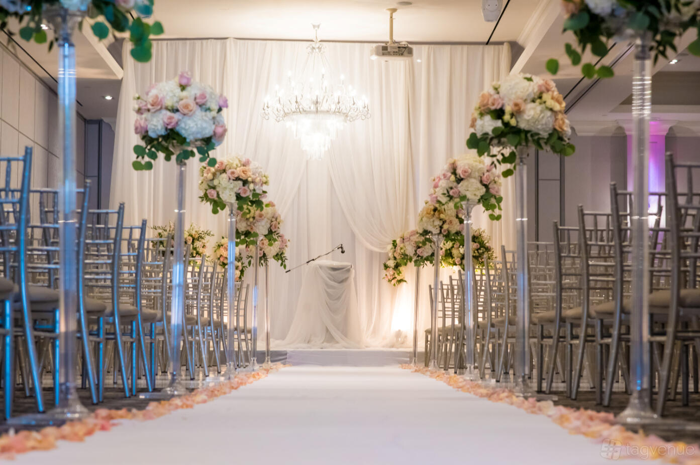 An event space with a chandelier, aisle lined with flower arrangements, and rows of silver chairs at Parkview Manor.