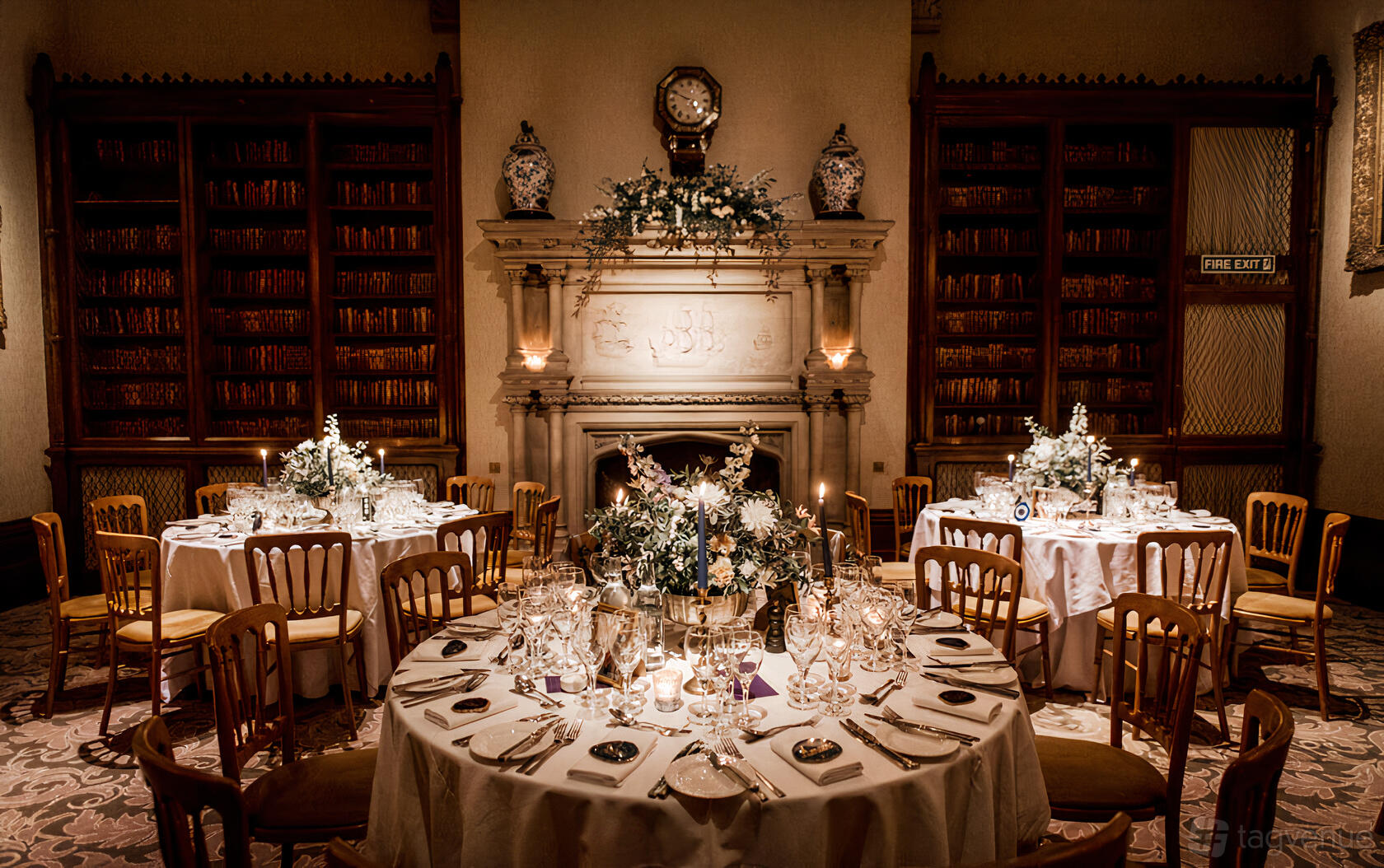 A banqueting hall with round tables, floral centerpieces, and a decorative fireplace flanked by bookshelves at The Elvetham.