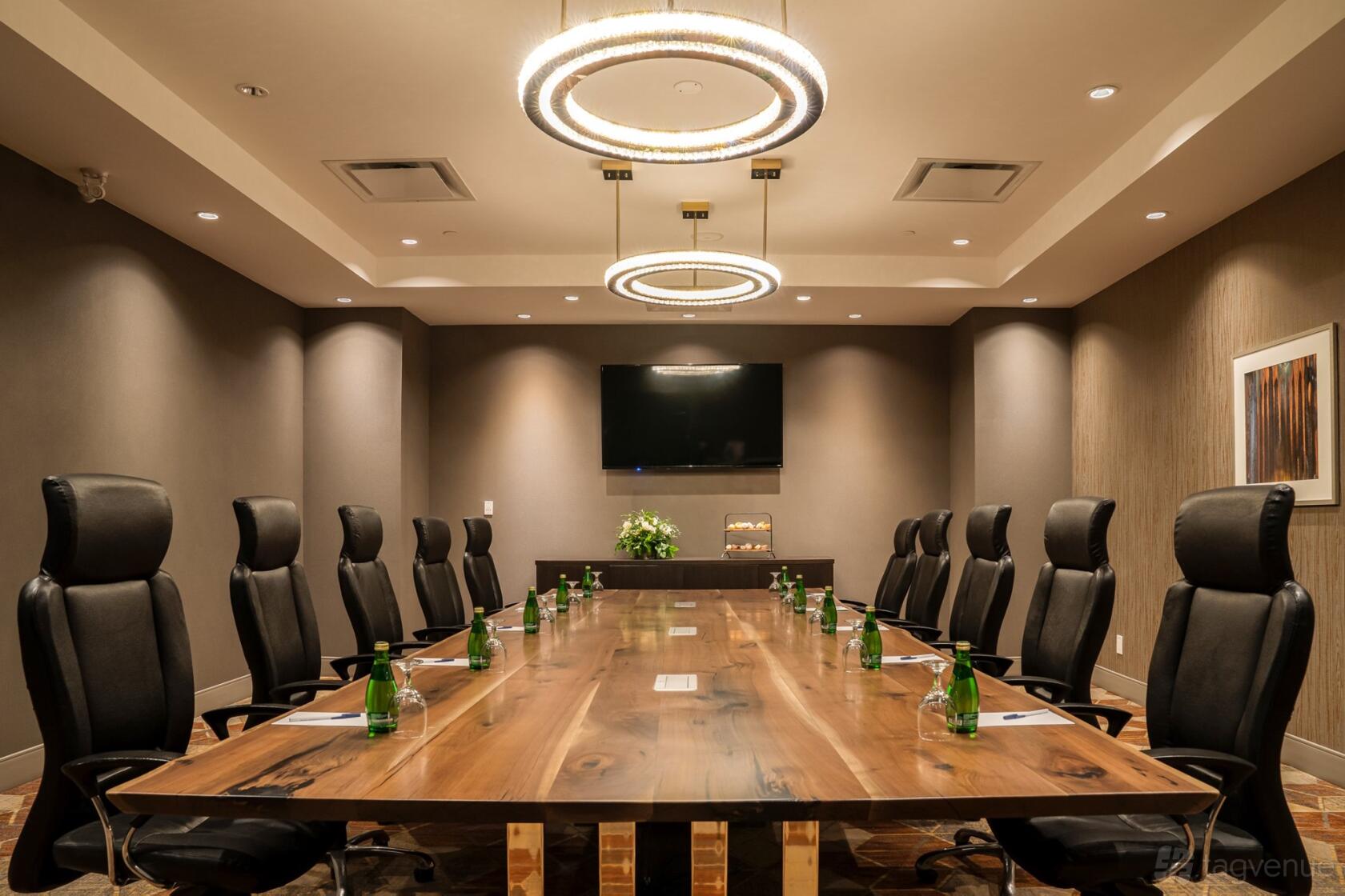 A hotel boardroom with a long wooden conference table, high-back chairs, modern ring chandeliers, and a wall-mounted TV at DoubleTree by Hilton Kitchener.