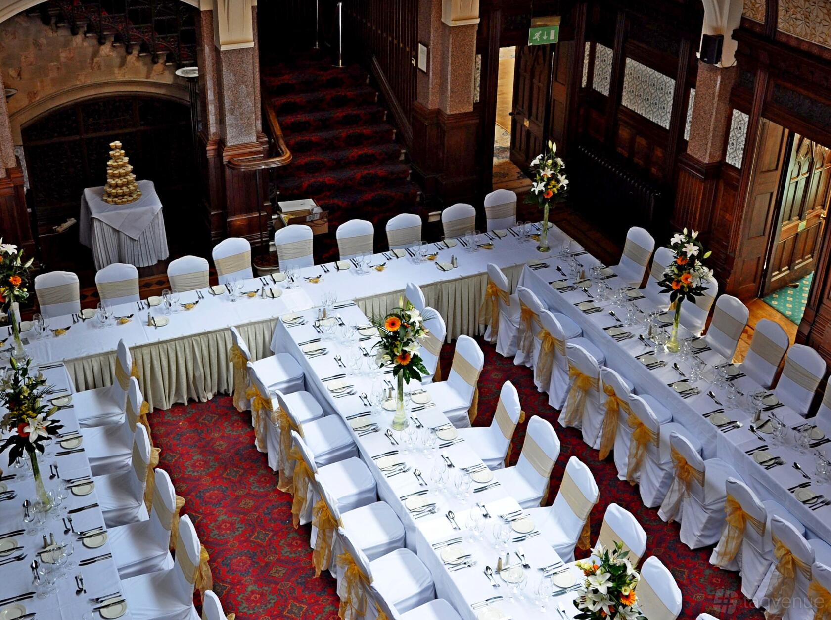 A hall with U-shaped banquet tables draped in white linens, gold chair sashes, and floral centerpieces at Highbury Hall.