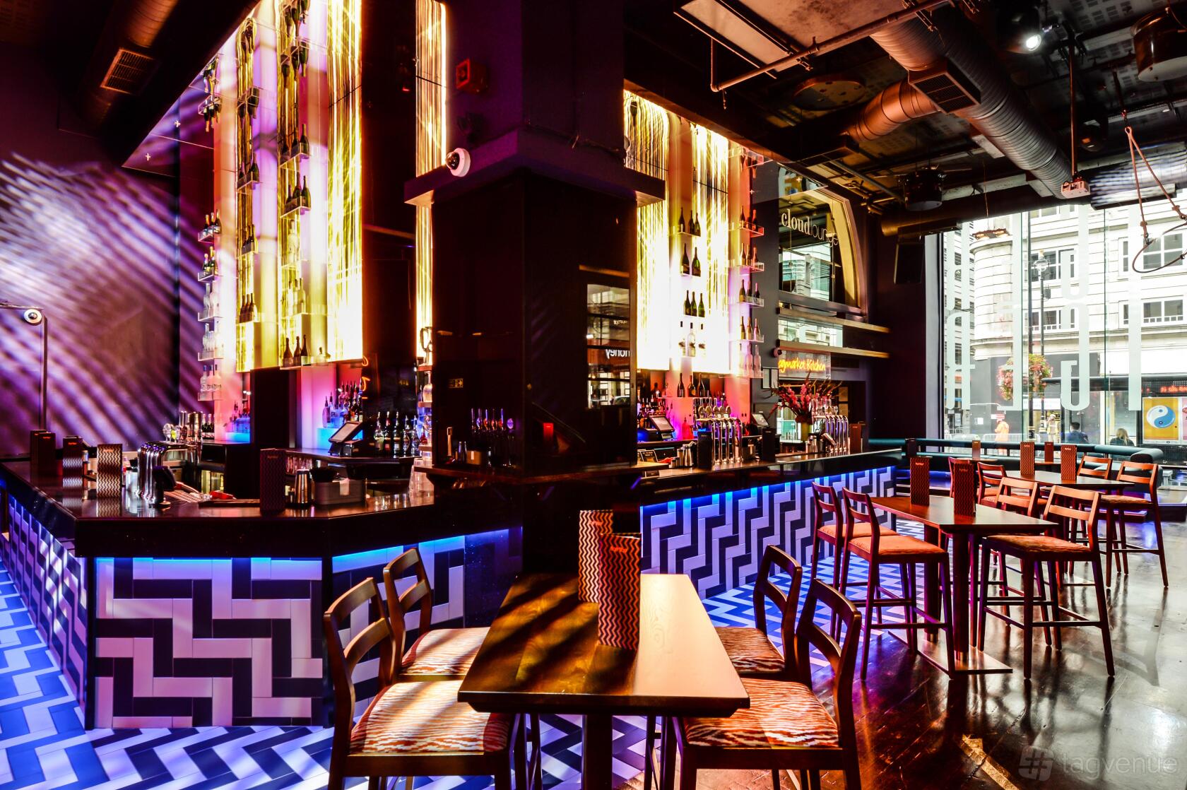 A nightclub with illuminated bar, zigzag patterned floor, tall tables, and colorful lighting at LUXE.