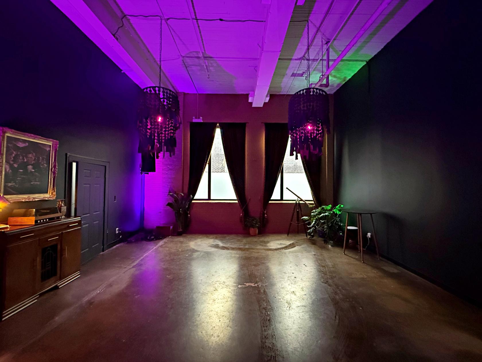 An event venue with dark walls, two chandeliers, large windows with drapes, and accent lighting at Roll This Way.