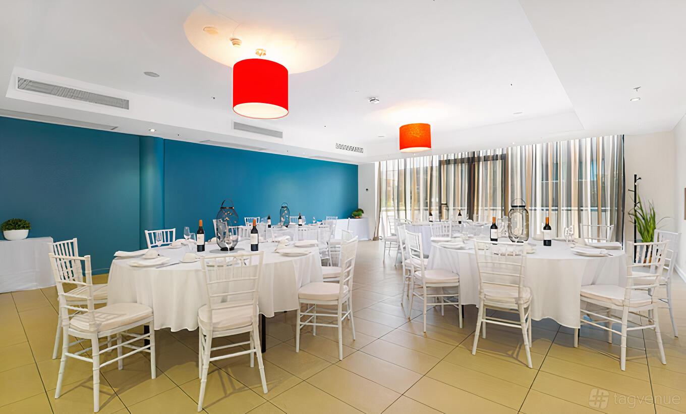 A hotel dining room with round tables draped in white linens, white chairs, and red pendant lamps at Sage Hotel Wollongong.