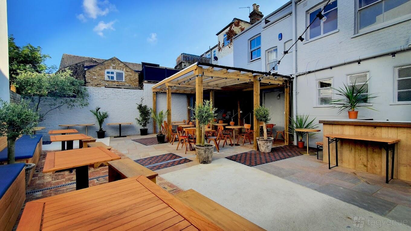 An outdoor pub space with wooden pergola, potted plants, high tables, and string lights at The Crossing Pub.
