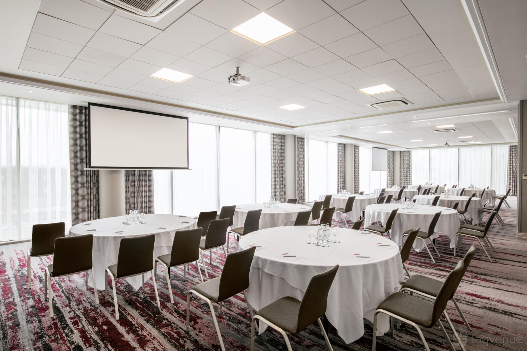 An event space with round tables covered in white linens, brown chairs, and floor-to-ceiling windows at Crowne Plaza London Docklands.